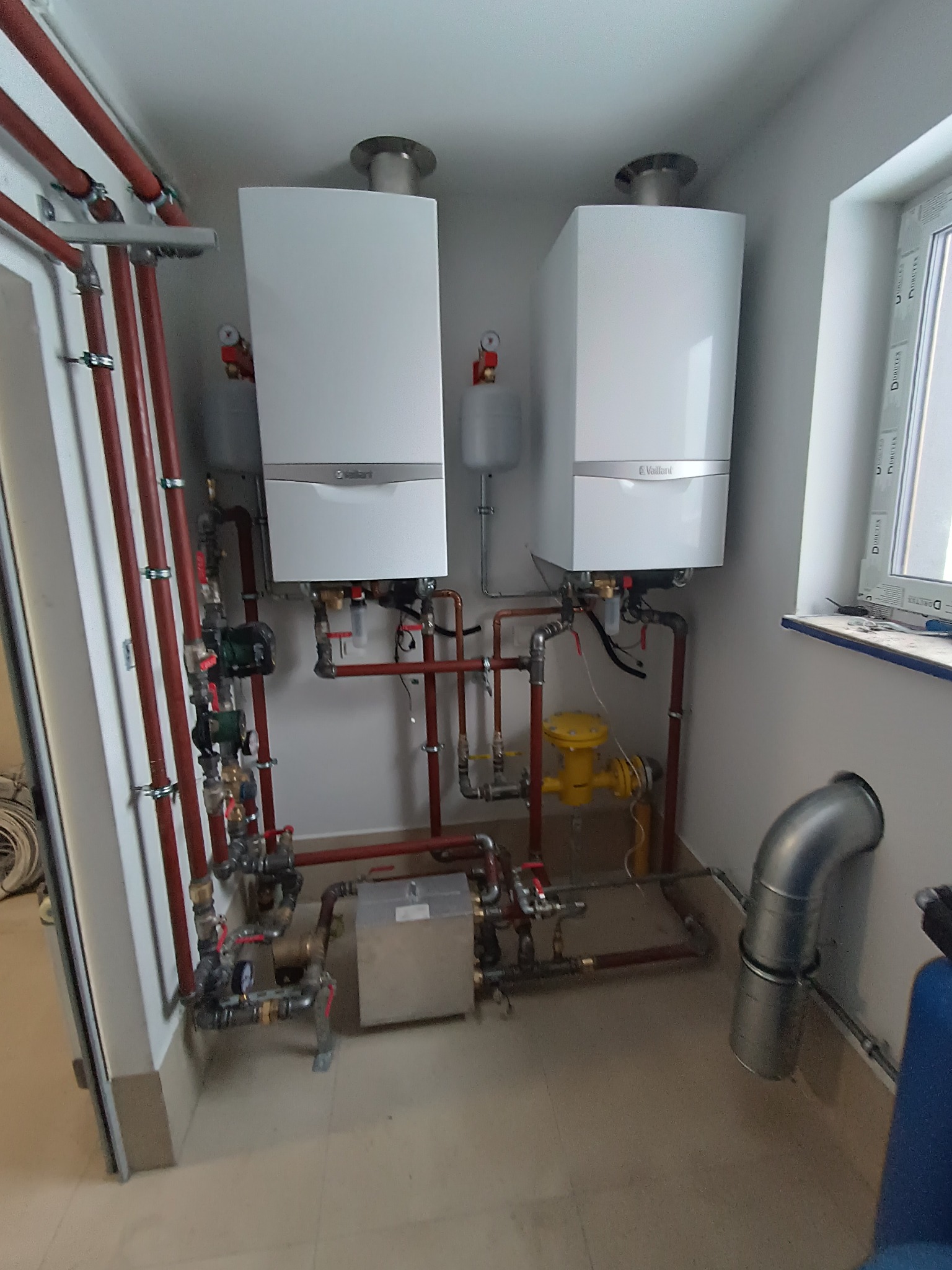 Dwa białe piece gazowe Vaillant zamontowane na ścianie w pomieszczeniu technicznym, z widocznymi rurami miedzianymi, zaworami i żółtym gazomierzem, oraz fragment okna po prawej stronie.