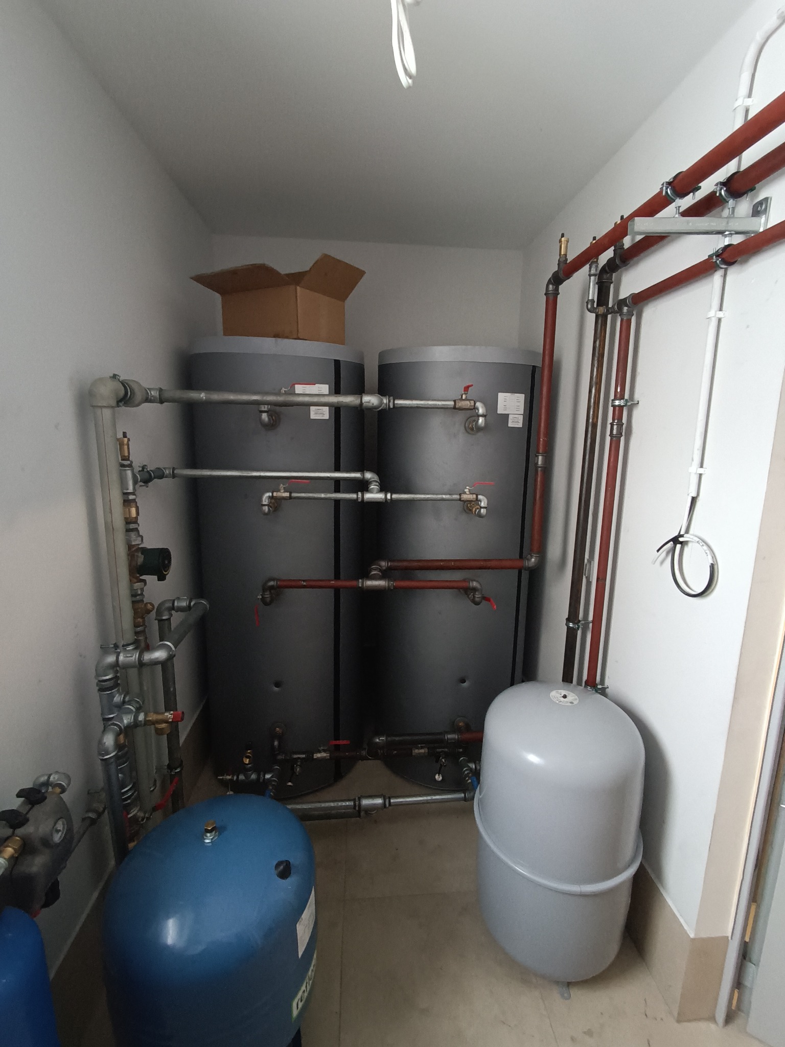 Dwa szare zbiorniki akumulacyjne ciepłej wody w kotłowni, z widocznymi rurami miedzianymi i stalowymi, zaworami, naczyniami przeponowymi w kolorze niebieskim i szarym oraz kartonem na jednym ze...