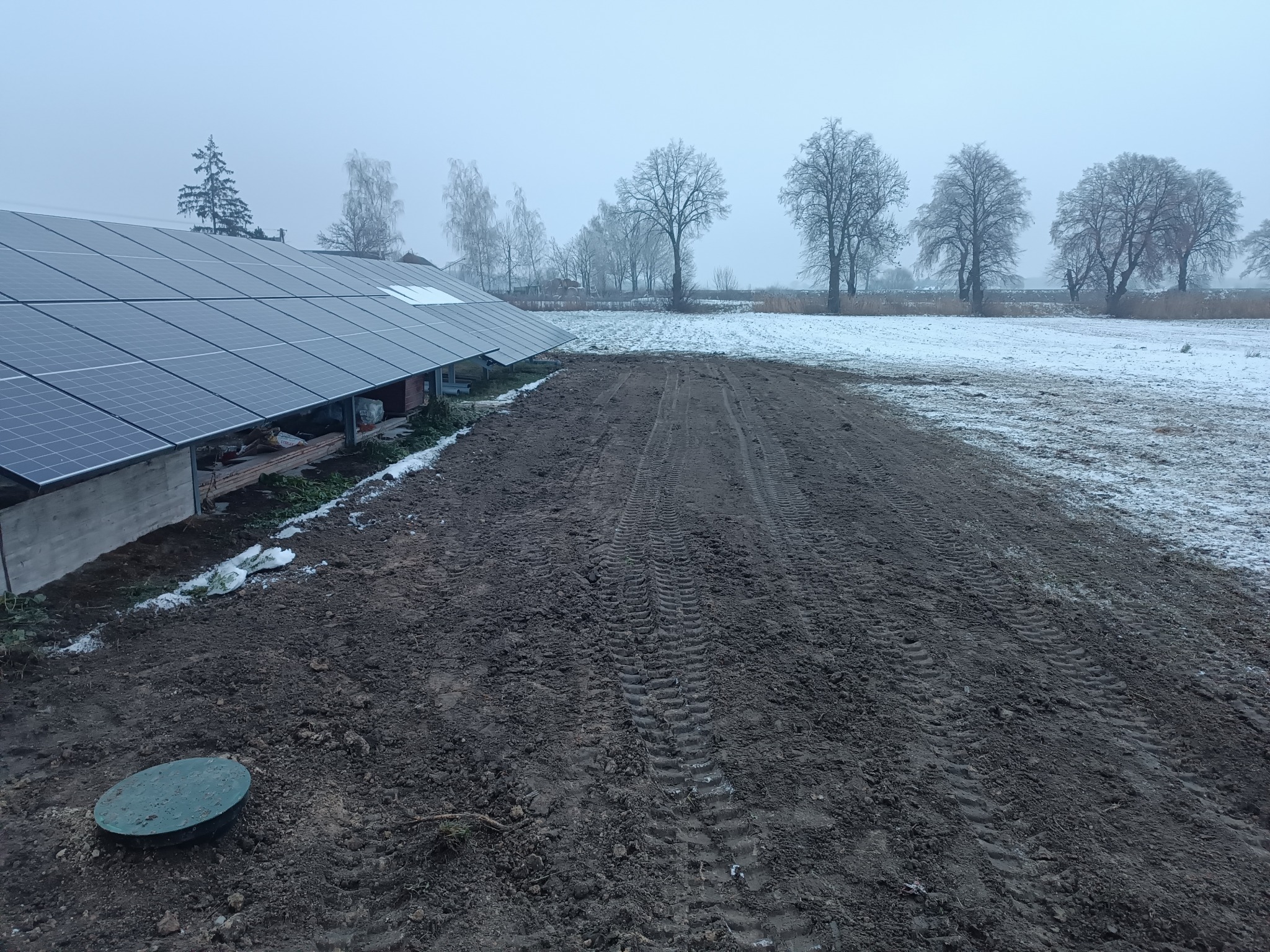 Instalacja paneli fotowoltaicznych na gruncie, widoczne ślady opon po sprzęcie budowlanym i pokrywa studzienki kanalizacyjnej w ziemi, w tle zaśnieżone drzewa i mgła.