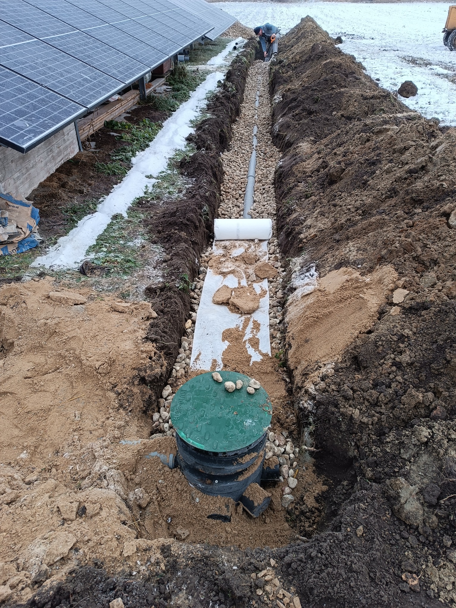 Wykopany rów z ułożonymi rurami drenarskimi i studzienką rewizyjną z zieloną pokrywą, zasypany częściowo żwirem; w tle panele solarne i pracownik przy dalszej części instalacji.