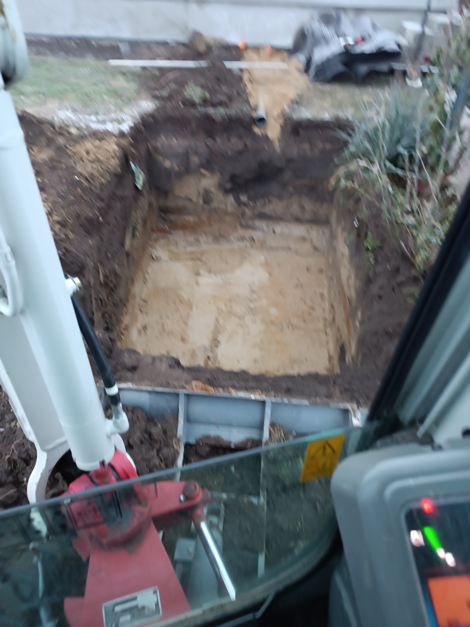 Widok z kabiny minikoparki na wykopany dół w ziemi, zabezpieczony szalunkiem, z widoczną instalacją rurową na dnie.