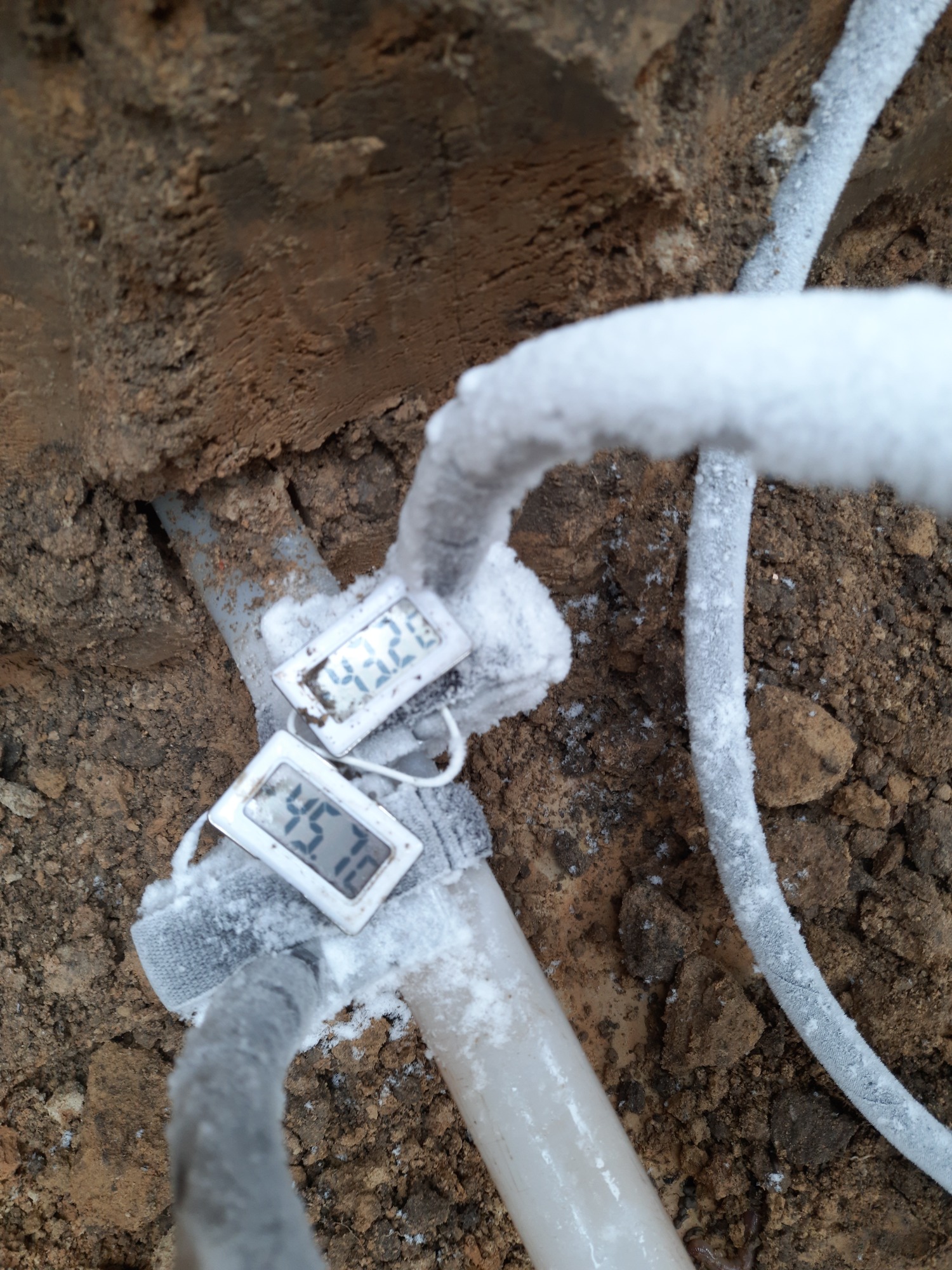 Zamarznięte rury w wykopie z podpiętymi termometrami wskazującymi temperaturę, ziemia wokół pokryta szronem.