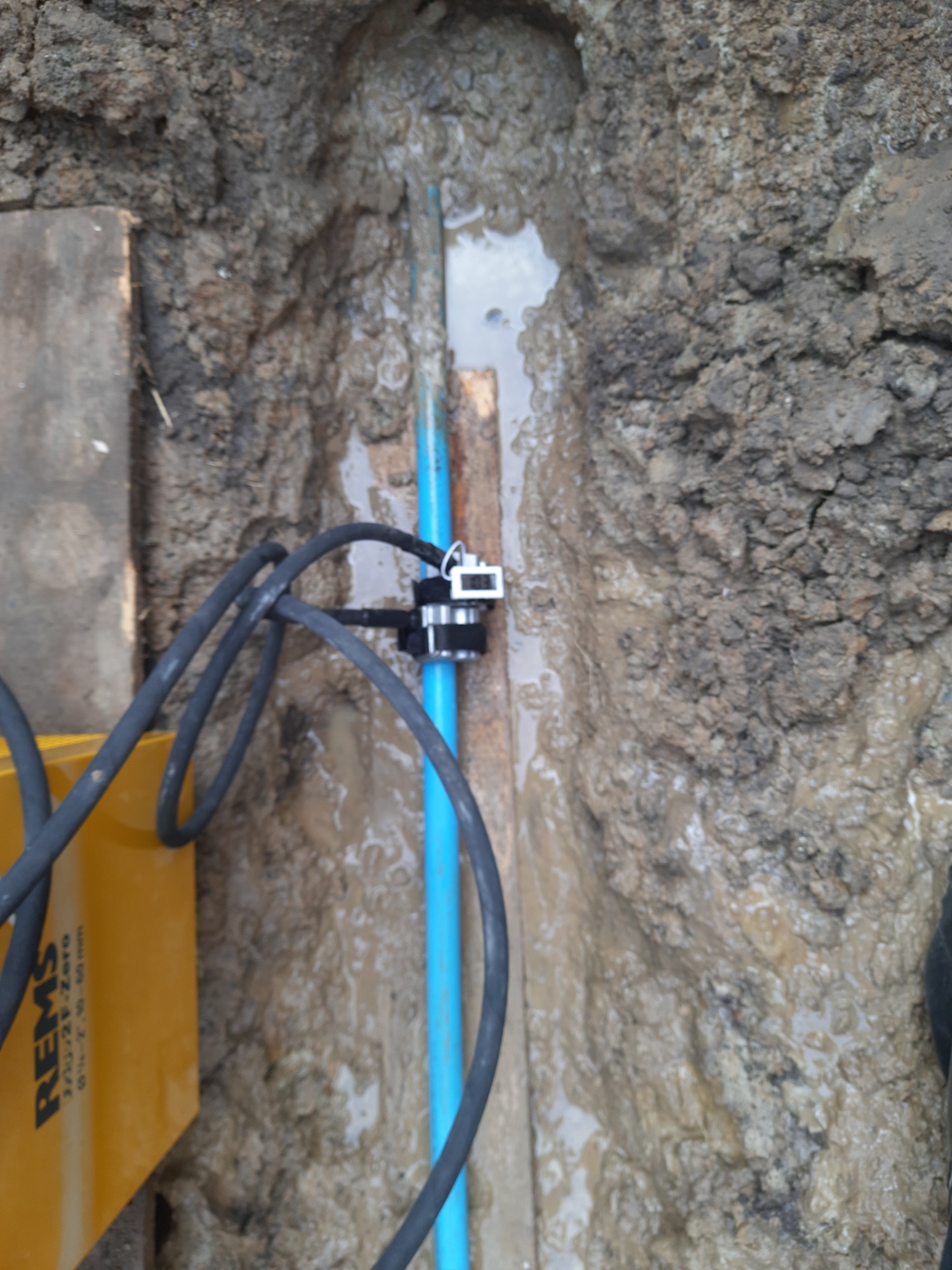 Błękitna rura wodociągowa w wykopie wypełnionym wodą, podłączona do czujnika, z widocznym fragmentem żółtej maszyny REMS i deską zabezpieczającą skarpę wykopu.