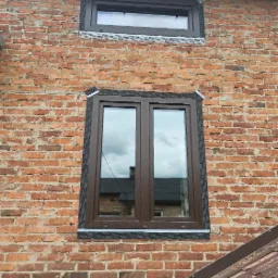 Dwa brązowe okna z PCV zamontowane w ścianie z czerwonej cegły, z widoczną taśmą uszczelniającą wokół ram okiennych, jedno okno mniejsze u góry i większe okno dwuskrzydłowe poniżej.