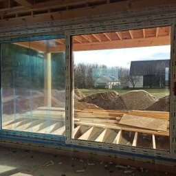 Windeys Roman Romaniuk - Montaż okna PCV w surowym stanie budynku. Widoczne drewniane elementy konstrukcyjne i folia ochronna na szybie. W tle krajobraz z budynkami.