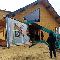 Windeys Roman Romaniuk - Montaż okna panoramicznego w domu szkieletowym przy użyciu dźwigu ssącego. Trzech monterów w trakcie pracy. Widoczne elementy konstrukcji budynku.