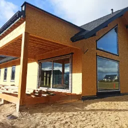 Dom w stanie surowym zamkniętym z zamontowanymi oknami i drzwiami przesuwnymi. Widoczna drewniana konstrukcja tarasu i elewacja z płyt OSB.