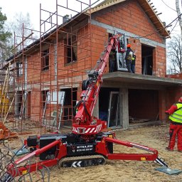 Windeys Roman Romaniuk - Montaż okna w surowym budynku z czerwonej cegły przy użyciu specjalistycznego, czerwonego dźwigu na gąsienicach; pracownicy w kaskach nadzorują proces.