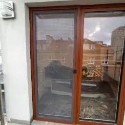 Brązowe przesuwne drzwi balkonowe z moskitierą, widok na balkon z zabezpieczonymi folią meblami, w tle budynki miejskie.