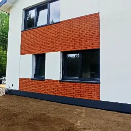 Fragment elewacji domu z białym tynkiem i wstawką z cegły klinkierowej, w której osadzone są czarne, nowoczesne okna.