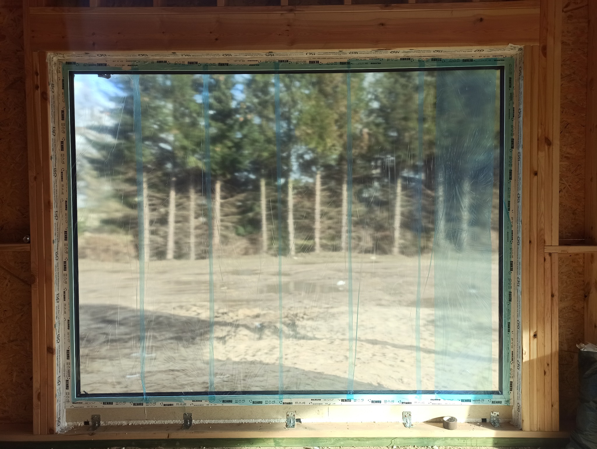 Nowe okno PCV marki Rehau, zabezpieczone folią ochronną, w surowym drewnianym otoczeniu. Widok na zewnątrz z zalesionym terenem w tle. Detal montażowy.