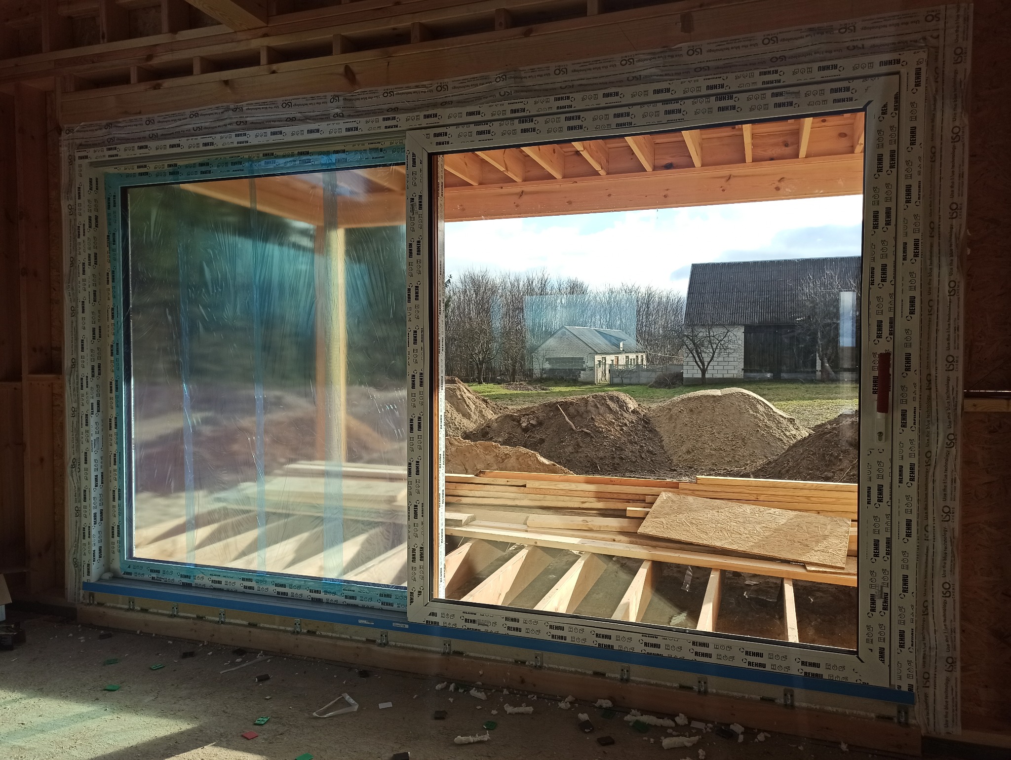 Montaż okna PCV w surowym stanie budynku. Widoczne drewniane elementy konstrukcyjne i folia ochronna na szybie. W tle krajobraz z budynkami.
