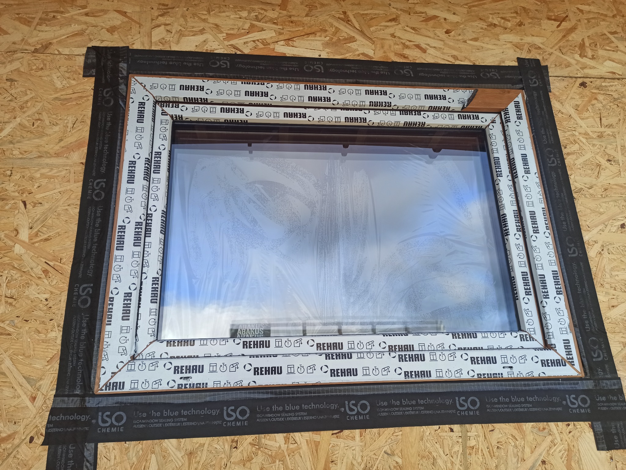Montaż okna PCV marki Rehau z folią ochronną i taśmą uszczelniającą ISO Chemie na surowej płycie OSB. Widoczne logo Rehau na ramie okiennej.