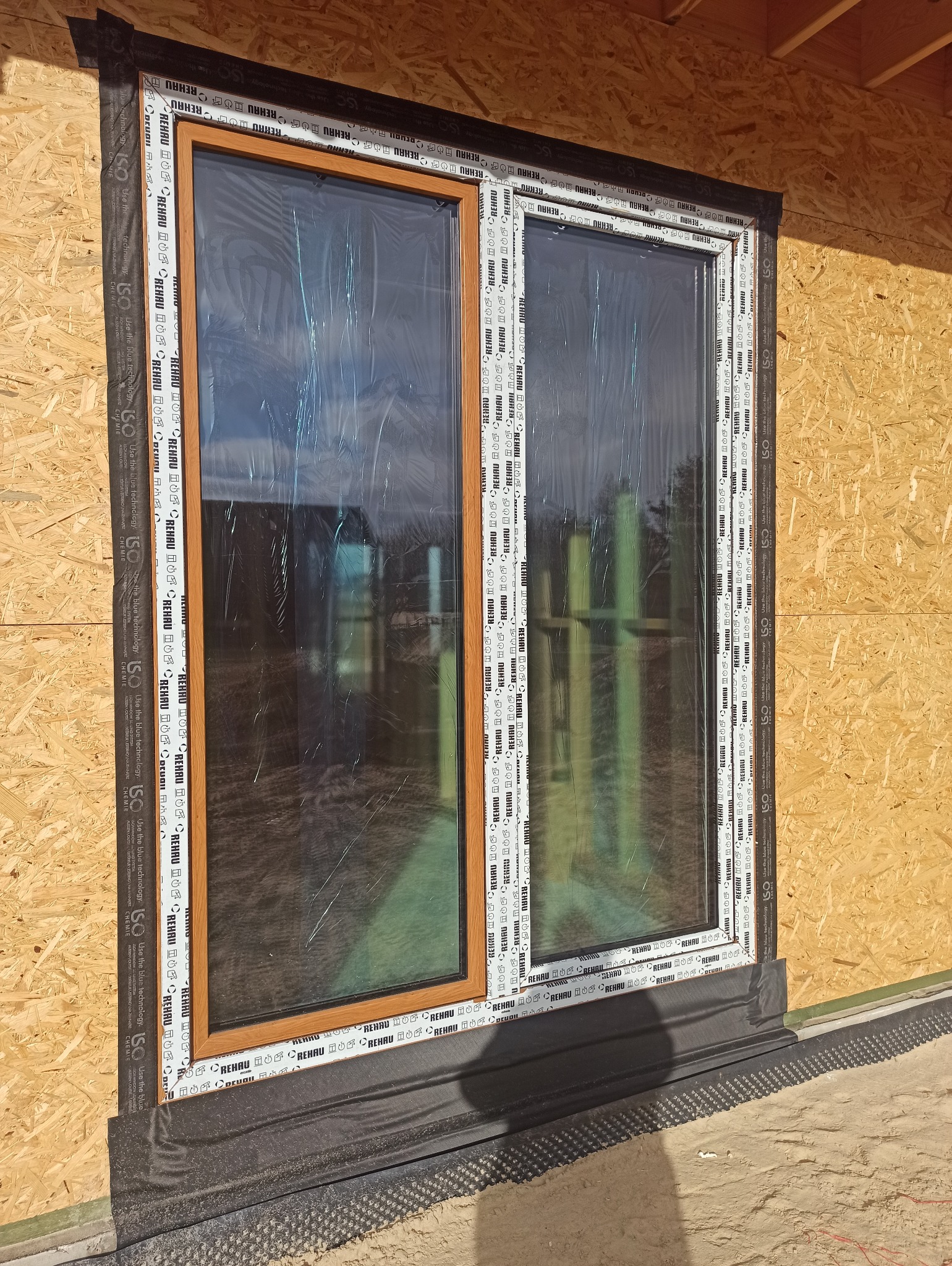 Montaż okna z drewnianą ramą, zabezpieczonego folią, w surowej ścianie z płyt OSB. Widoczne taśmy uszczelniające i folia hydroizolacyjna.