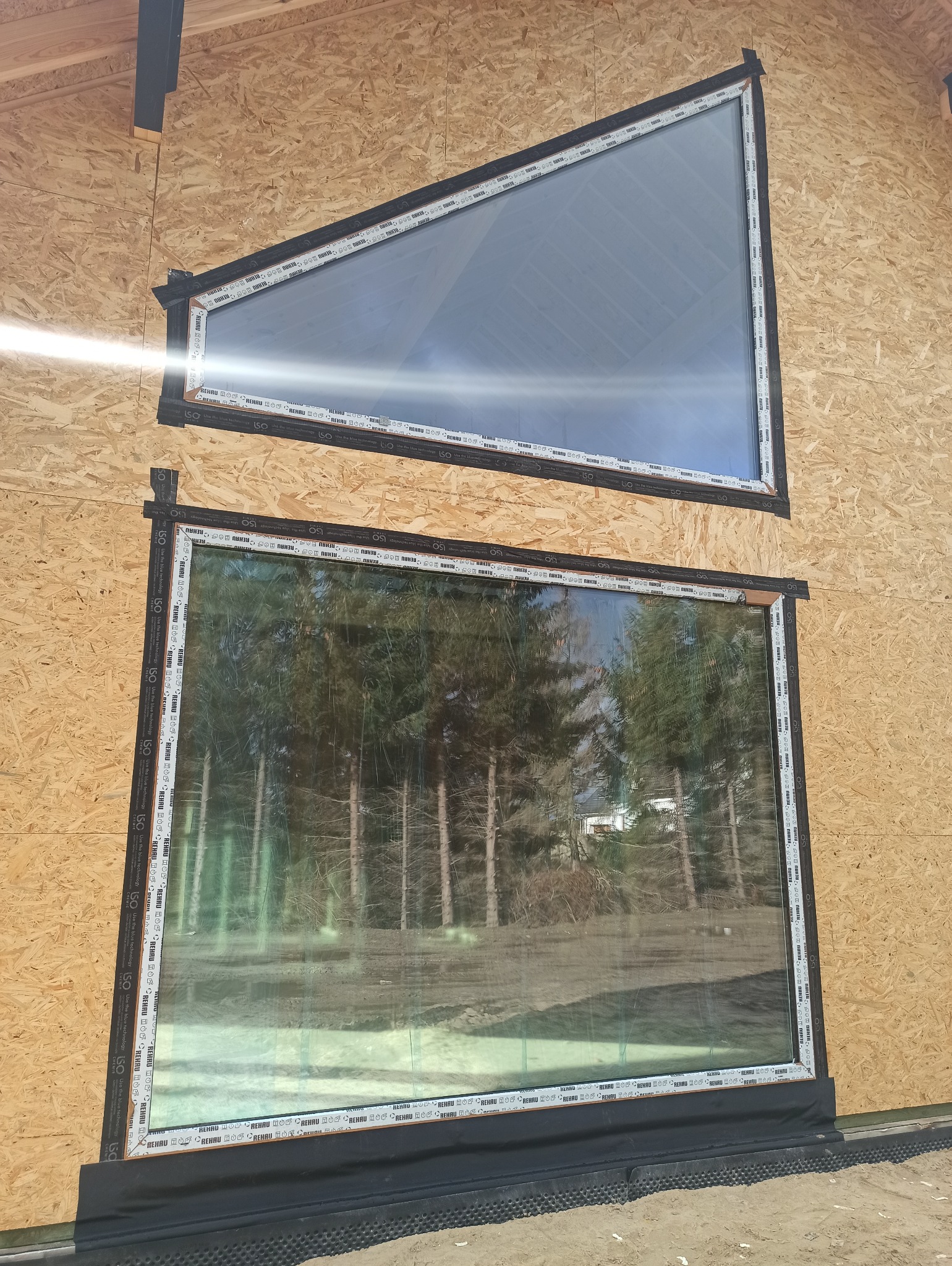 Montaż okien o nietypowych kształtach: trójkątne okno nad prostokątnym, oba z ramami Rehau, w surowym wnętrzu z płyty OSB. Widok na las przez dolne okno.