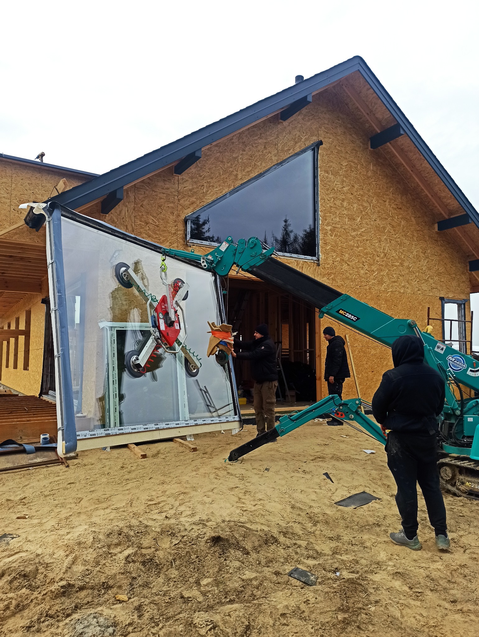 Montaż okna panoramicznego w domu szkieletowym przy użyciu dźwigu ssącego. Trzech monterów w trakcie pracy. Widoczne elementy konstrukcji budynku.