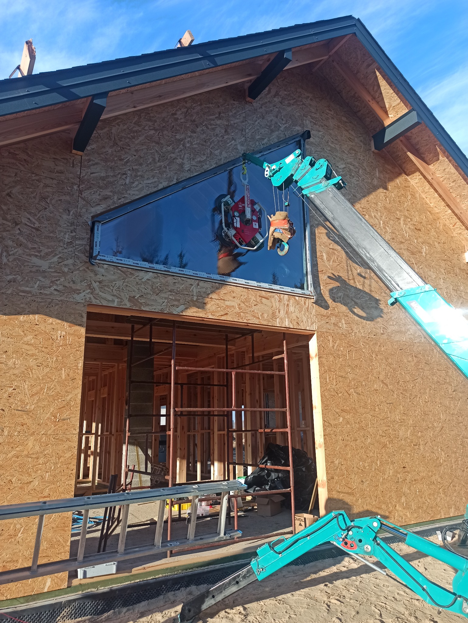 Montaż okna o nietypowym kształcie na elewacji budynku w stanie surowym. Użycie specjalistycznego dźwigu do przenoszenia szyby. Widoczne rusztowania i drabina.