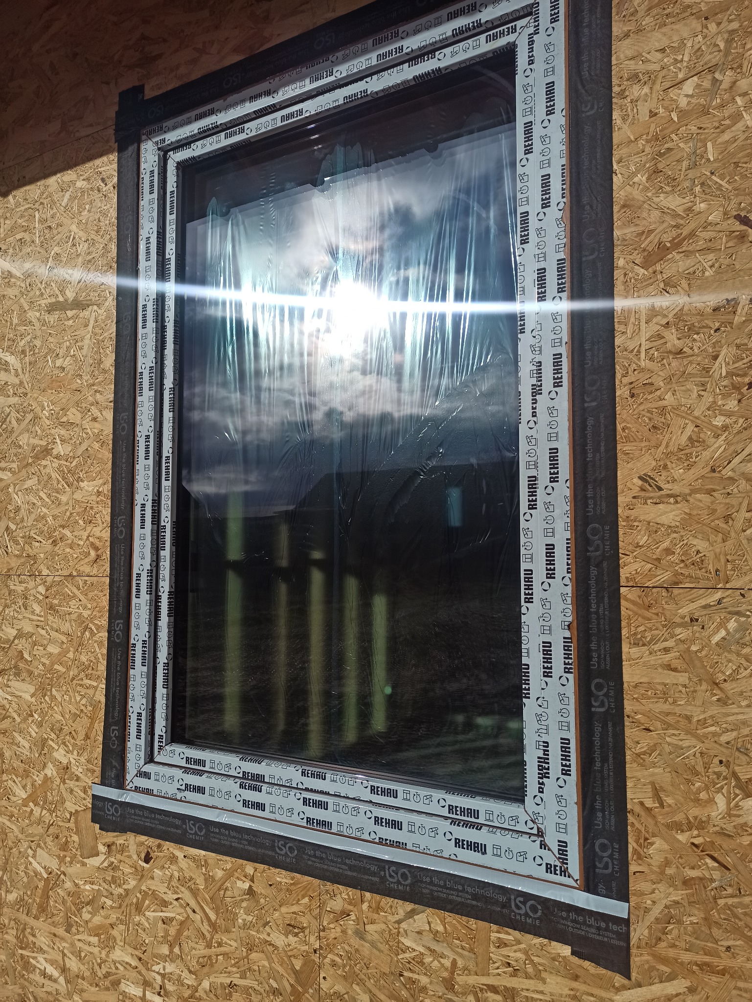 Okno PCV Rehau w trakcie montażu, zabezpieczone folią ochronną, widoczne uszczelki i taśmy izolacyjne, surowa płyta OSB w tle. Detal montażowy.