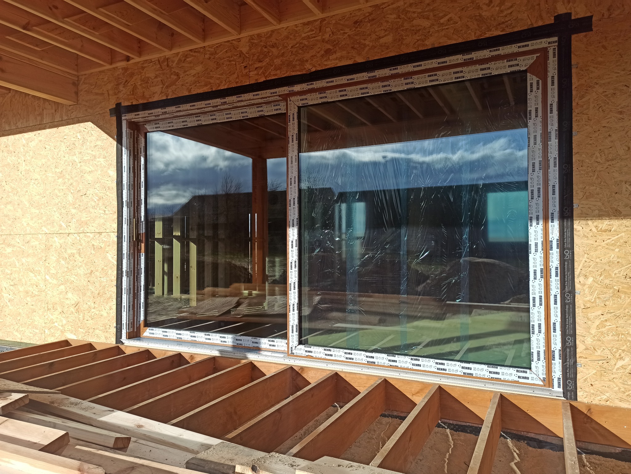 Montaż okna przesuwnego z brązową ramą w surowym stanie budynku. Widoczne drewniane elementy konstrukcyjne i folia ochronna na szybach. Ujęcie z dołu.