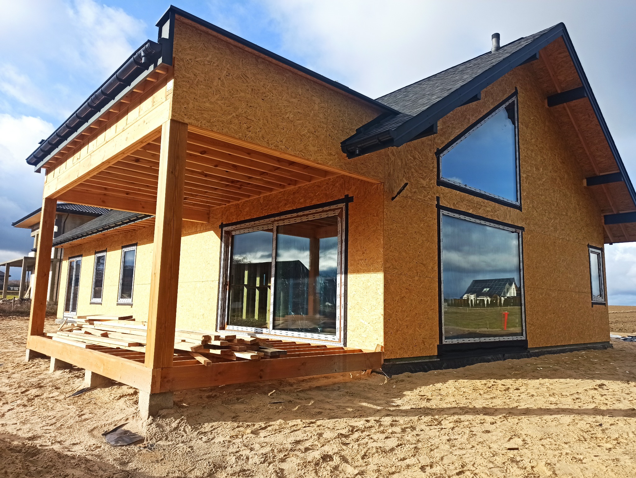 Dom w stanie surowym zamkniętym z zamontowanymi oknami i drzwiami przesuwnymi. Widoczna drewniana konstrukcja tarasu i elewacja z płyt OSB.