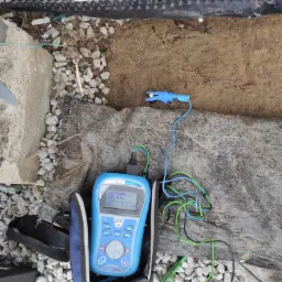 Miernik rezystancji uziomu Eurotest MI 3125, z widocznymi przewodami pomiarowymi i sondą, umieszczony na geowłókninie i żwirze w wykopie przy fundamencie budynku.