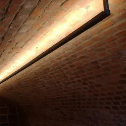 Oświetlenie LED zamontowane na metalowej szynie pod ceglanym, łukowym sufitem piwnicy, z widoczną drabiną w tle.