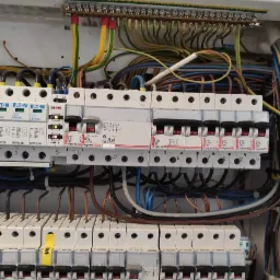 Otwarta skrzynka elektryczna z widocznymi bezpiecznikami Legrand i Eaton, okablowaniem oraz listwą zaciskową u góry, z żółto-zielonymi przewodami ochronnymi.