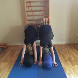 Dwie osoby w pozycji psa z głową w dół, używające drabinki gimnastycznej z czerwonymi linami i poduszek do ćwiczeń na niebieskich matach.