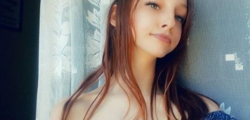 Portret młodej kobiety z długimi rudymi włosami, stojącej przy oknie z zasłoną w kwiaty i niebieską ścianą w paski.