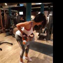 Kobieta w sportowym stroju wykonuje ćwiczenie wiosłowania z dwoma kettlebells w siłowni, z widocznym sprzętem do ćwiczeń w tle.