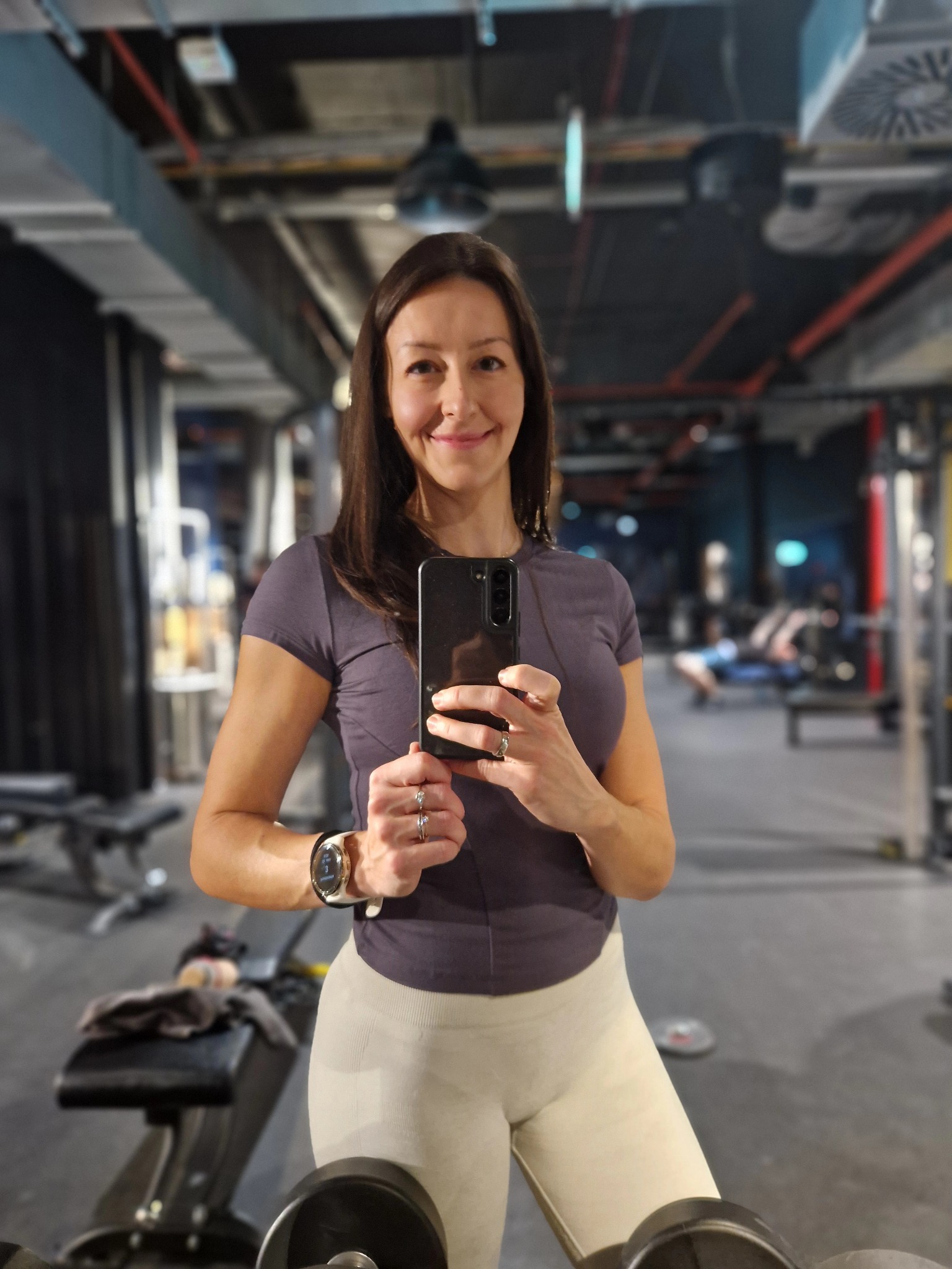 Uśmiechnięta kobieta w sportowym stroju robi selfie w siłowni, widoczny zegarek sportowy i pierścionki, w tle sprzęt do ćwiczeń i ćwiczący ludzie.