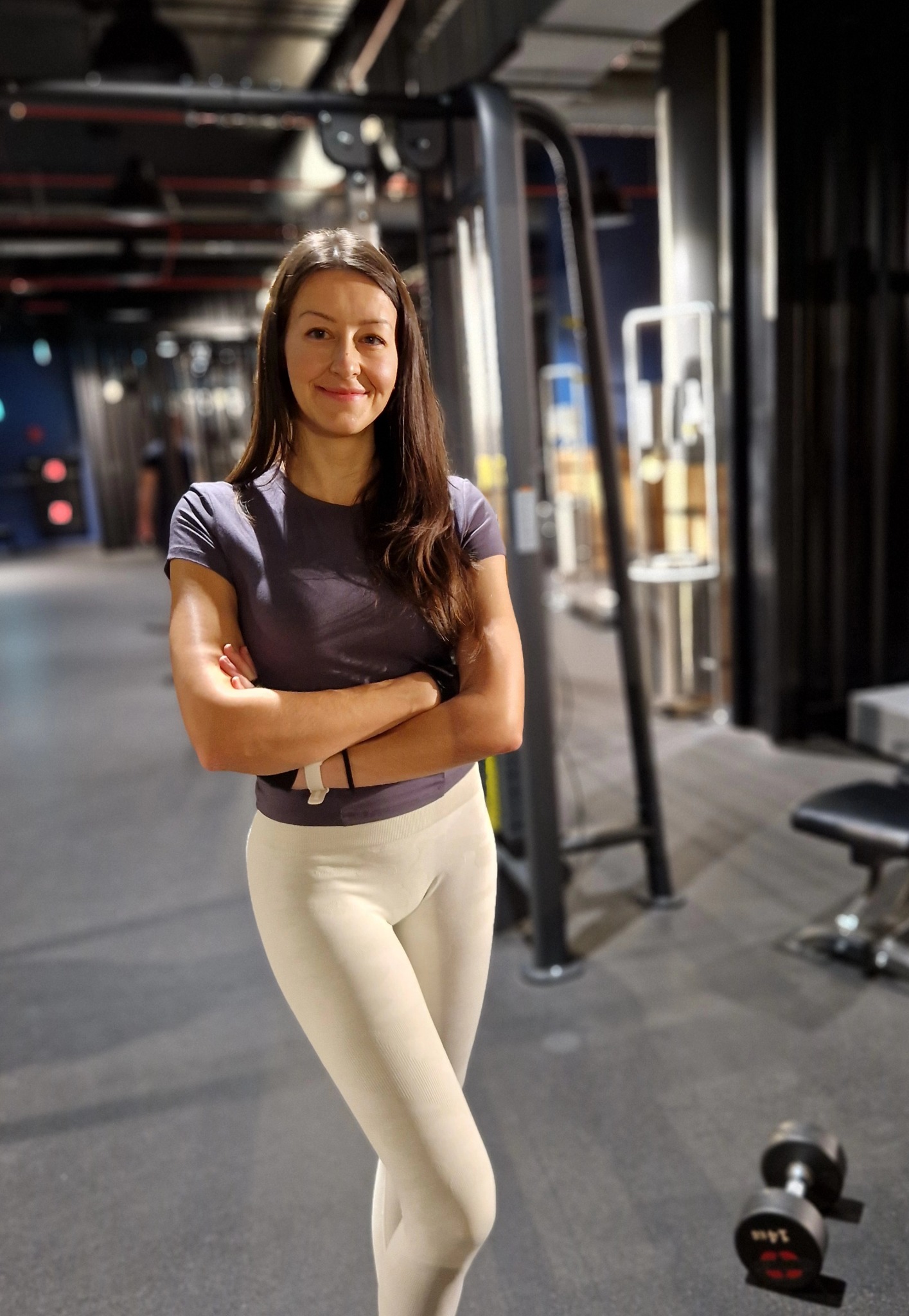 Uśmiechnięta kobieta w sportowym stroju, z założonymi rękami, stoi na tle siłowni z maszynami i hantlami. Dobre oświetlenie podkreśla jej sylwetkę.