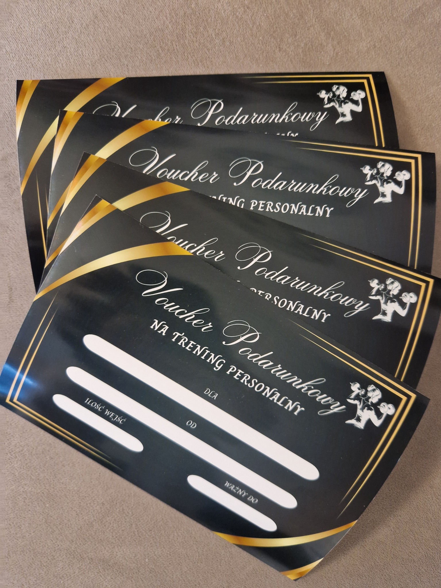 Stos czarnych voucherów podarunkowych na trening personalny z złotymi akcentami i sylwetką osoby ćwiczącej z hantlami, leżących na beżowym tle.