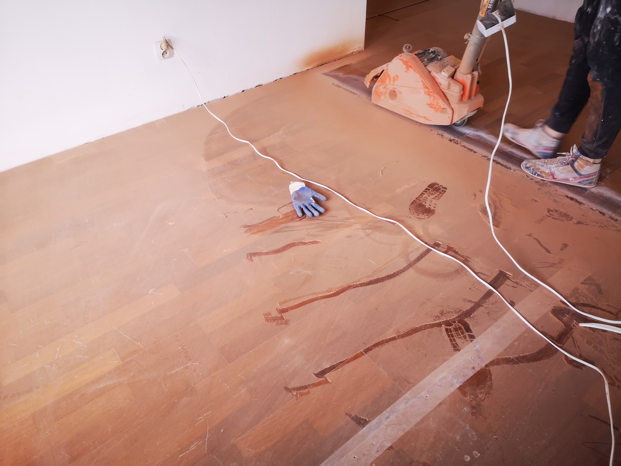 Szlifowanie drewnianej podłogi za pomocą pomarańczowej cykliniarki, widoczne ślady obróbki, biały kabel i niebieska rękawica robocza na podłodze.