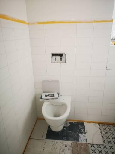 Biała toaleta podwieszana, w trakcie montażu, z otwartym pudełkiem na desce sedesowej, widoczne białe płytki na ścianach z żółtą taśmą malarską wzdłuż sufitu i częściowo położona podłoga z różnymi...