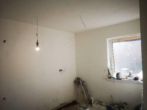 Pokój w trakcie remontu: surowe ściany, prowizoryczne oświetlenie żarówką na kablu, okno z widocznymi śladami prac budowlanych i narzędzia na podłodze.