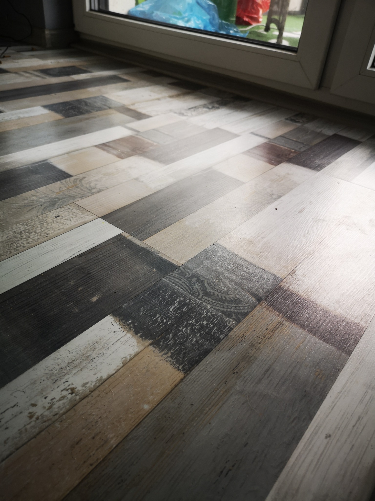 Ułożone pod kątem, różnokolorowe panele podłogowe imitujące drewno, z widoczną strukturą i wzorem, przy białej ramie okiennej.