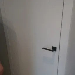 Białe drzwi wewnętrzne z czarną, kwadratową klamką, widoczne trzy pionowe frezy na skrzydle drzwiowym.