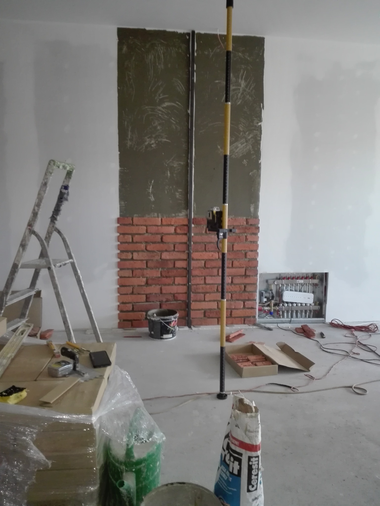Ściana w trakcie wykańczania z fragmentem pokrytym cegłą dekoracyjną, górna część przygotowana pod gładź, widoczna drabina, poziomica laserowa, materiały budowlane i instalacja ogrzewania podłogowego.