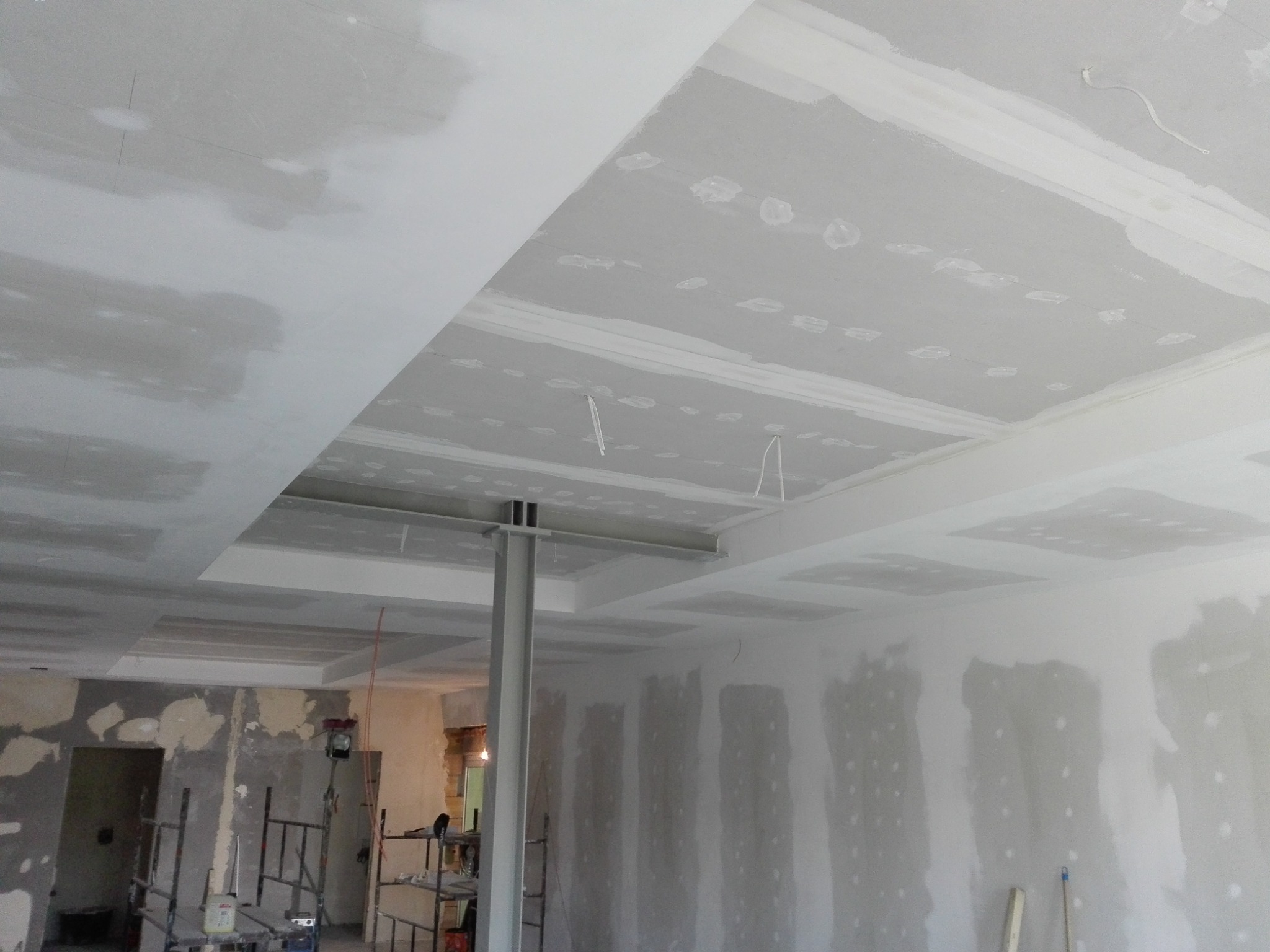 Sufit podwieszany z płyt kartonowo-gipsowych w trakcie montażu, widoczne łączenia i szpachlowanie, metalowa konstrukcja nośna, w tle rusztowanie i ściany w trakcie przygotowania.