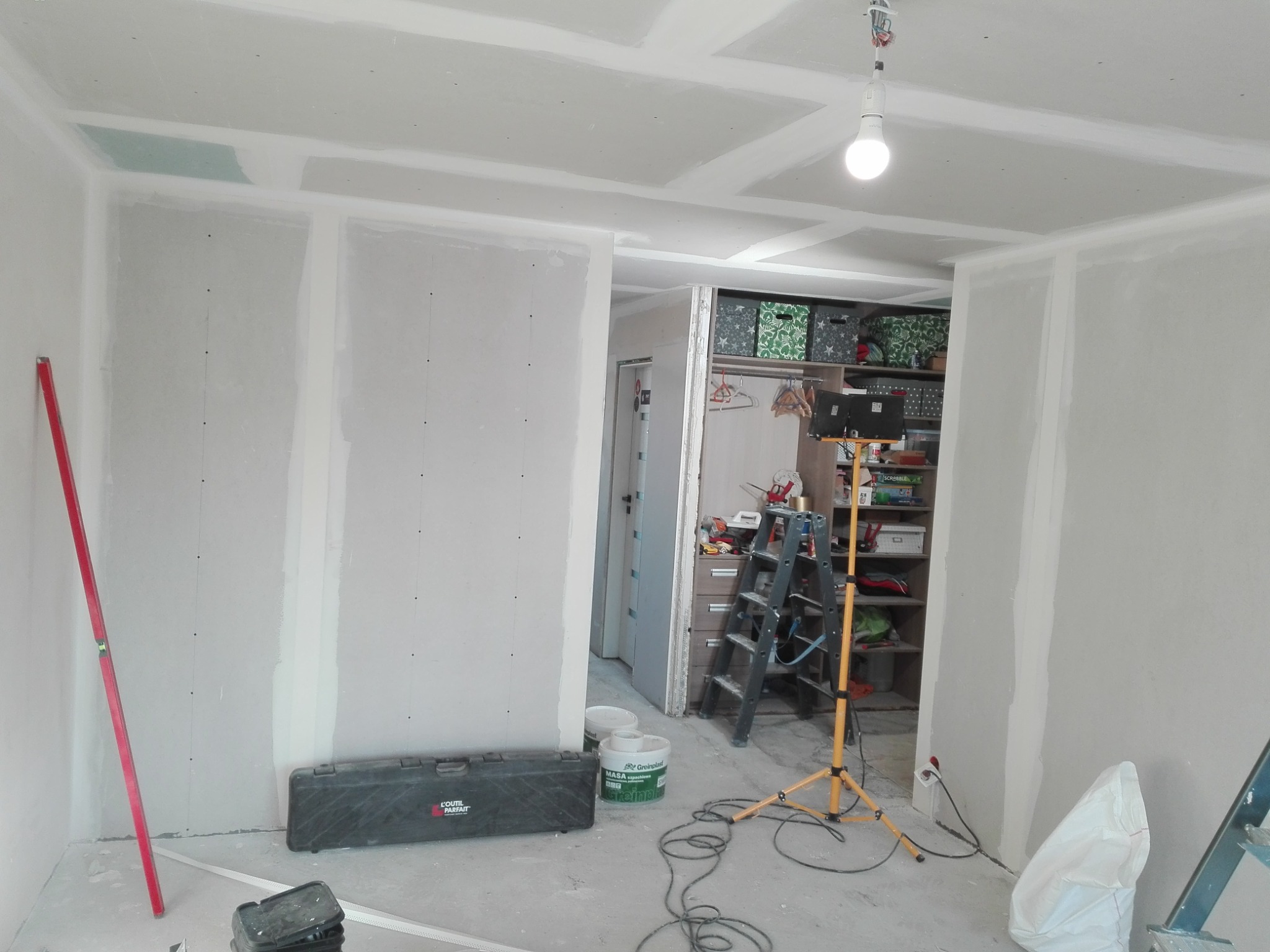 Wnętrze w trakcie montażu płyt kartonowo-gipsowych na ścianach i suficie, widoczny poziomica, skrzynka narzędziowa, drabina i lampa budowlana, na suficie widoczne łączenia płyt.