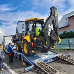 Żółto-czarna koparko-ładowarka MST 642 PLUS na niebieskiej platformie transportowej, przygotowywana do rozładunku przez dwóch mężczyzn na tle błękitnego nieba z chmurami.