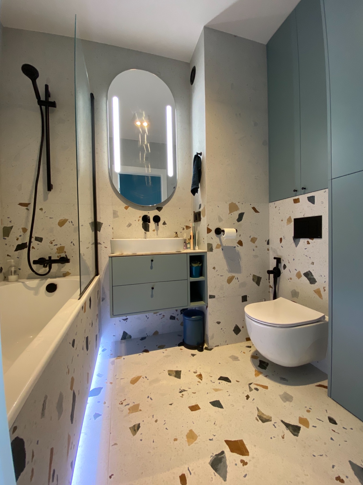 Nowoczesna łazienka z płytkami lastryko na ścianach i podłodze, podświetlana wanna, umywalka z szafką w kolorze pastelowej zieleni, owalne lustro z oświetleniem LED i czarna armatura.