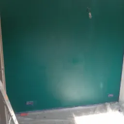 Świeżo pomalowana ściana w odcieniach zieleni z widocznymi śladami taśmy malarskiej przy listwach i gniazdkach, zabezpieczona folią podłoga i drabina malarska z boku.