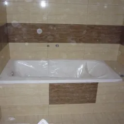 Nowa wanna, zabezpieczona folią ochronną, zabudowana płytkami w odcieniach beżu i brązu, zamontowana w wykończonej łazience.