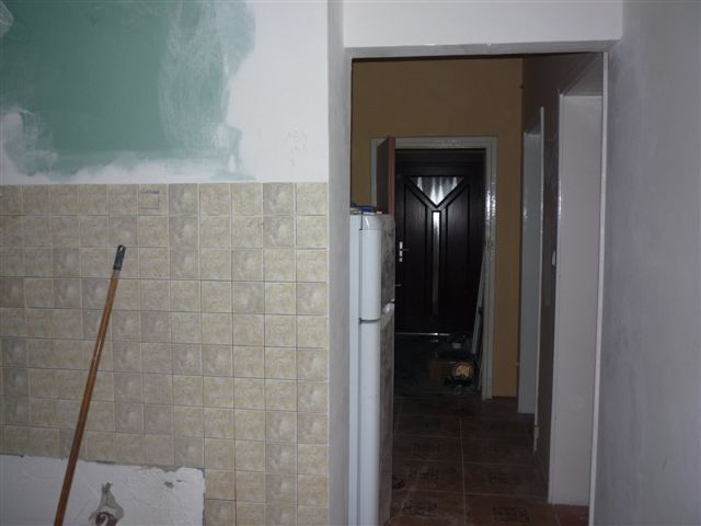 Wnętrze mieszkania w trakcie remontu: ściana z częściowo położonymi beżowymi płytkami, fragment pomalowanej na zielono ściany, widoczna lodówka i drzwi do innego pomieszczenia.