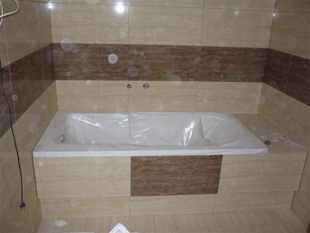 Nowa wanna, zabezpieczona folią ochronną, zabudowana płytkami w odcieniach beżu i brązu, zamontowana w wykończonej łazience.