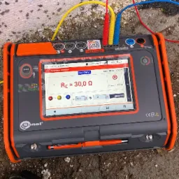 Miernik Sonel MPI-540 z podłączonymi przewodami pomiarowymi, wyświetlający wynik rezystancji uziemienia 30,0 &Omega; na ekranie dotykowym, umieszczony na szorstkiej, zabrudzonej powierzchni.