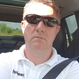 Ryszard Czerwiński - Mężczyzna w okularach przeciwsłonecznych, ubrany w białą koszulkę polo z logo Hummel, siedzi w samochodzie z zapiętym pasem bezpieczeństwa, widok przez szybę na zielone tereny.