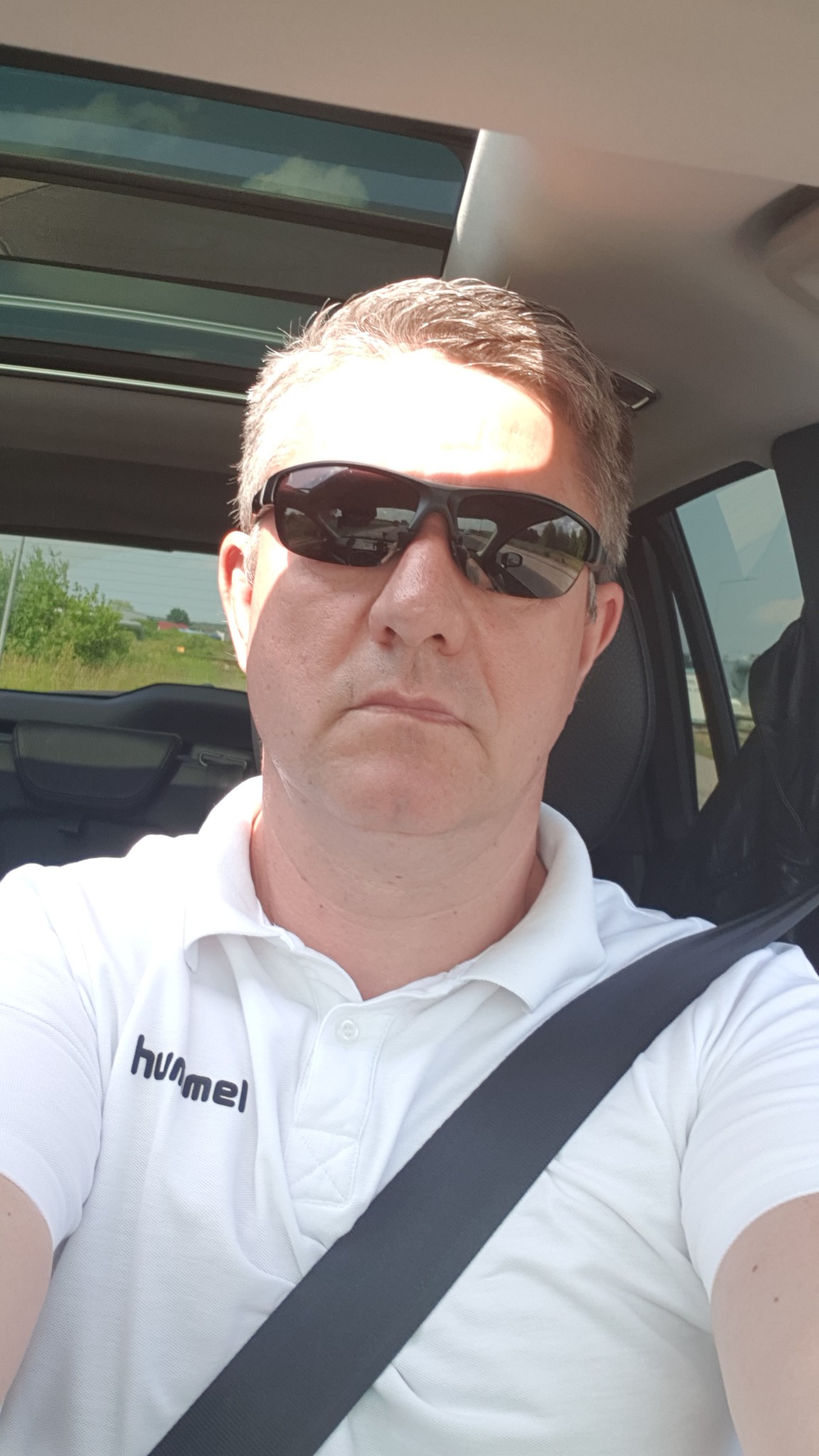 Mężczyzna w okularach przeciwsłonecznych, ubrany w białą koszulkę polo z logo Hummel, siedzi w samochodzie z zapiętym pasem bezpieczeństwa, widok przez szybę na zielone tereny.